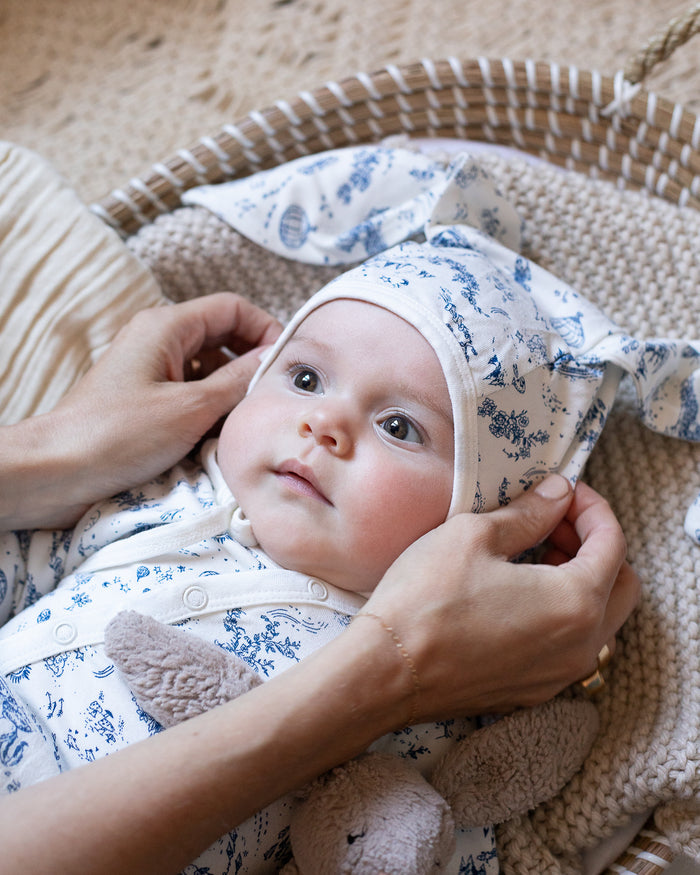 A baby wearing a blue and white floral bonnet with bunny ears, and a blue and white toile bodysuit.