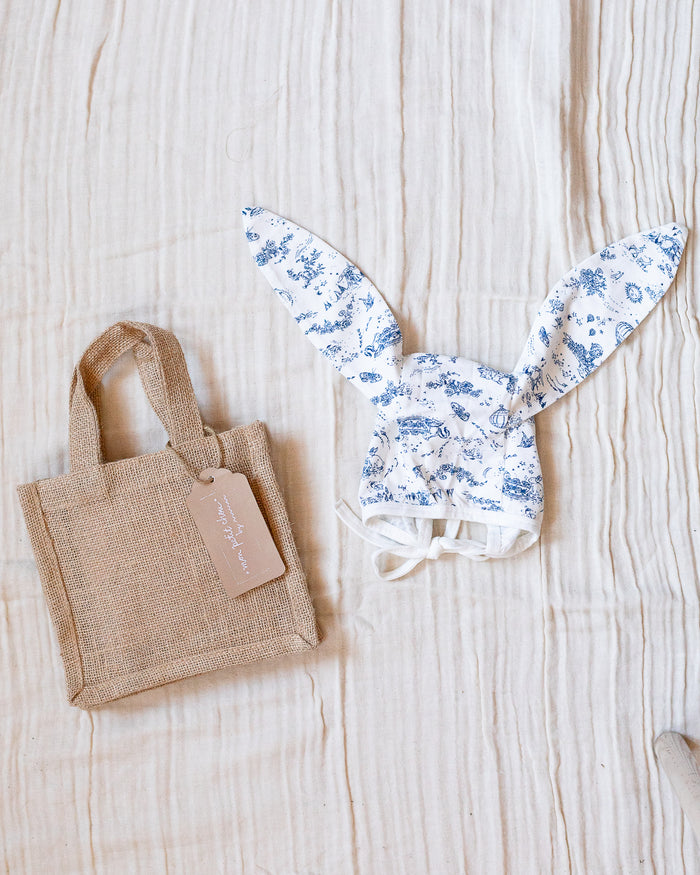 Blue and white toile bonnet with bunny ears next to a beige jute bag.