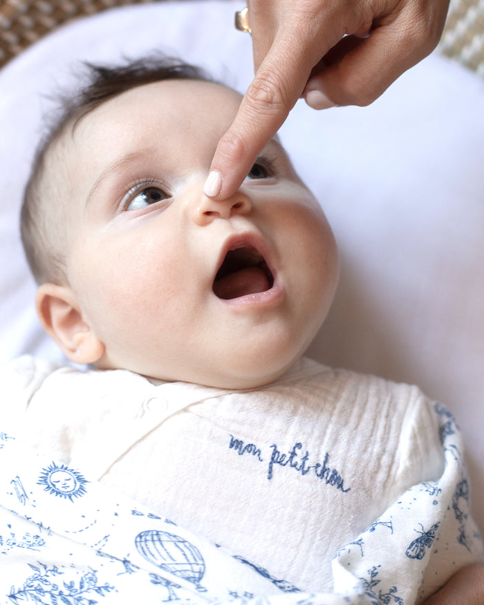Baby wearing a soft white muslin bodysuit with "mon petit chou" embroidered in blue on the chest, meaning "my little darling" in French.
