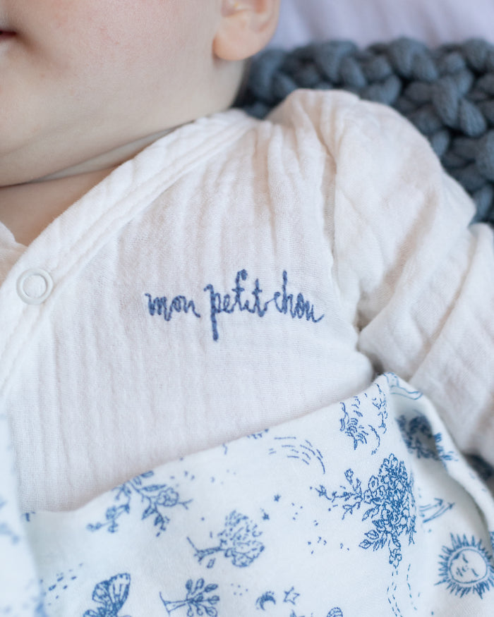 Baby wearing a soft white muslin bodysuit with "mon petit chou" embroidered in blue on the chest, wrapped in a blue-and-white toile blanket.
