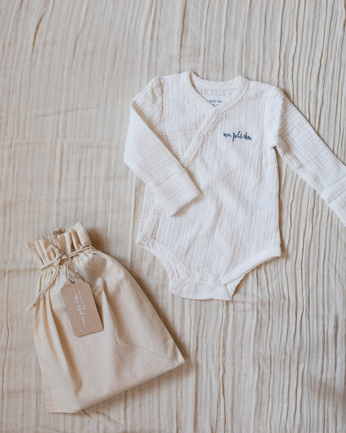 White muslin bodysuit with "mon petit chou" ("my little darling" in French) embroidered in blue on the chest, placed next to a small beige toile bag.