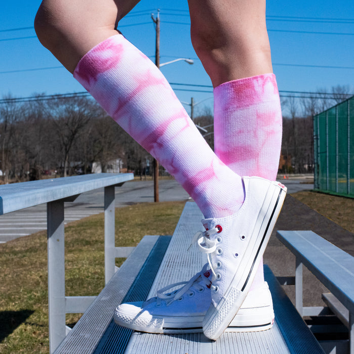 Knee-high compression socks with a pink tie-dye pattern worn with white sneakers, standing on a bench outdoors.