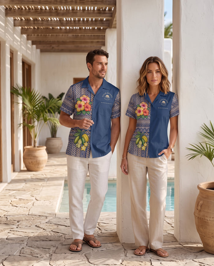 A man and a woman wearing matching blue short-sleeve camp shirts with floral and geometric patterns, standing in a sunny outdoor patio corridor next to a pool.