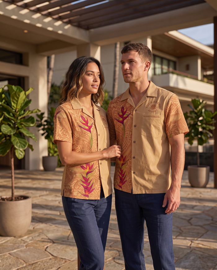 A man and a woman standing outdoors in front of a modern house, wearing matching tan short-sleeve camp-collar shirts with a tropical floral print in pink and green, paired with navy pants.