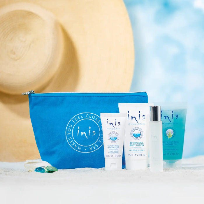 A blue Inis pouch with four skincare products arranged in front of it on a white surface, with a large straw hat and a soft blue water-themed backdrop in the background.