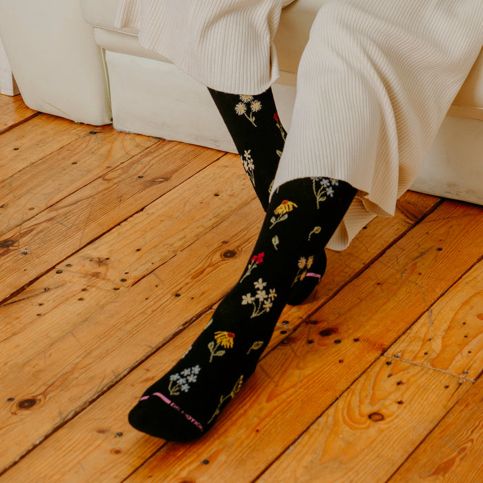 Knee-high black compression socks with colorful wildflower patterns, worn with light-colored pants on wooden floor.