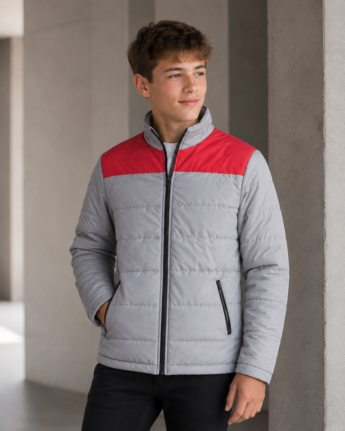 A young man wearing a light gray puffer jacket with a contrasting red yoke and black zipper details, standing in a modern concrete corridor.